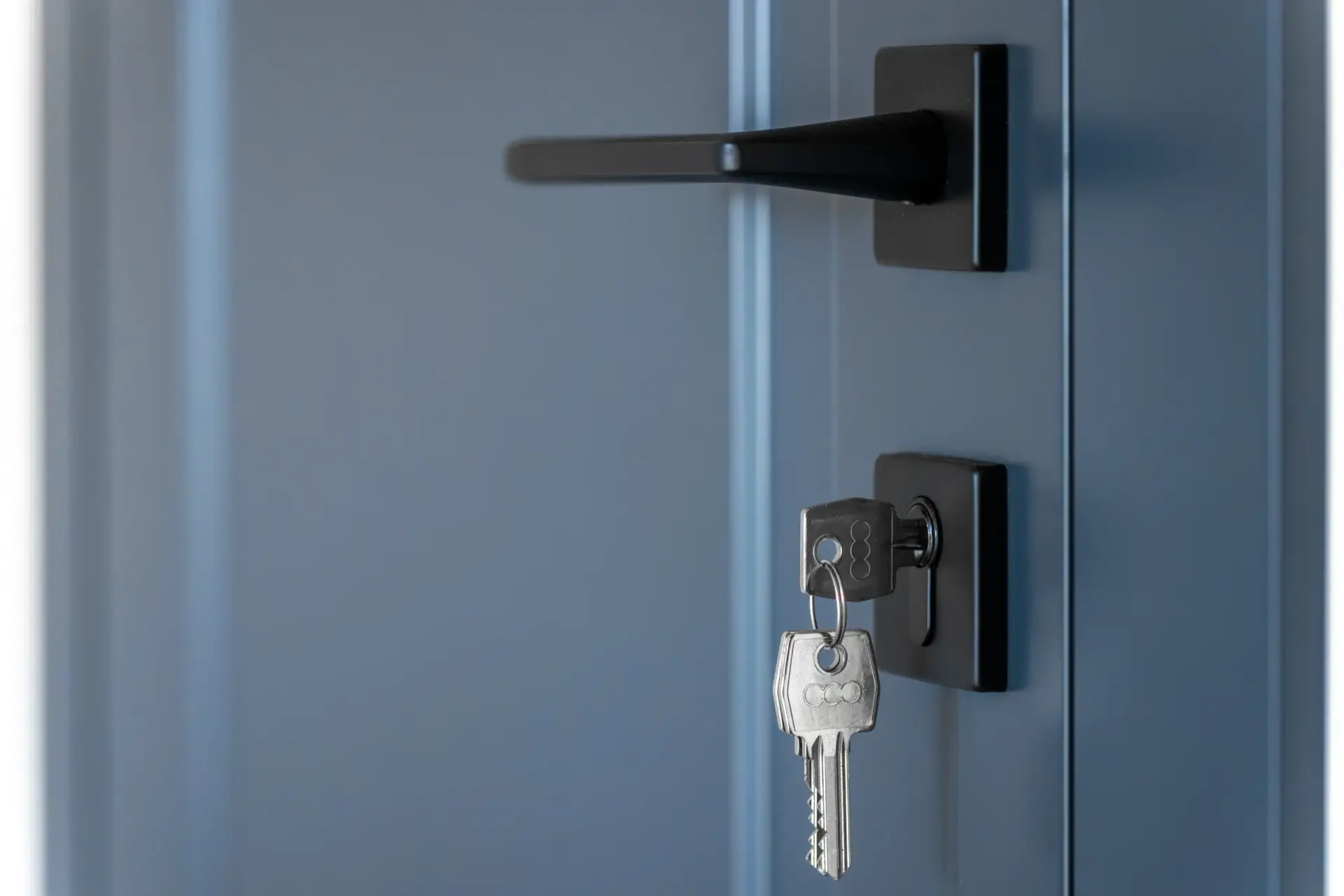 Close up of a modern matte black door handle and silver keys in the keyhole of a blue gray door panel.